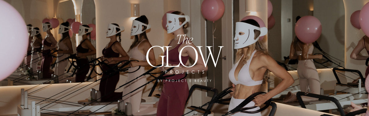 Women in Pilates class wearing LED red light therapy masks at The Glow Projects by Project E Beauty, stylish activewear in white, pink, and burgundy, with pink balloons in mirrored studio.